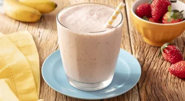 Fotografia em tons de rosa claro em uma bancada de madeira de cor marrom. Ao centro, um pires azul claro com um copo em cima contendo o frozen. Ao lado, há um pano amarelo e um recipiente contendo morangos espalhados. Ao fundo, há duas bananas. Fotografia em tons de rosa claro em uma bancada de madeira de cor marrom. Ao centro, um pires azul claro com um copo em cima contendo o frozen. Ao lado, há um pano amarelo e um recipiente contendo morangos espalhados. Ao fundo, há duas bananas.