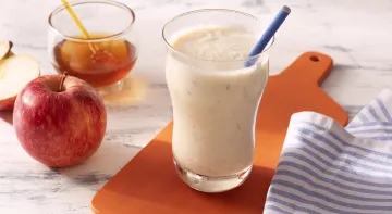 Fotografia em tons de branco em uma bancada de madeira de cor branca. Ao centro, uma tábua de madeira laranja com um copo em cima contendo a bebida. Ao lado, há um pano branco listrado, 1 maçã e um recipiente com mel. Fotografia em tons de branco em uma bancada de madeira de cor branca. Ao centro, uma tábua de madeira laranja com um copo em cima contendo a bebida. Ao lado, há um pano branco listrado, 1 maçã e um recipiente com mel.