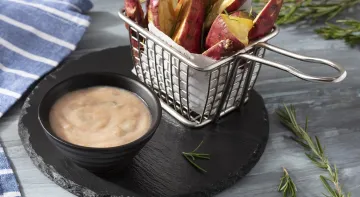 Fotografia em tons claros, em uma bancada de madeira cinza com um pano azul listrado. Ao centro, um prato redondo preto contendo um recipiente com o molho e um outro recipiente com as batatas rústicas. Ao fundo, alguns ramos de alecrim espalhados.