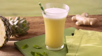 Fotografia em tons de amarelo em uma bancada de madeira de cor marrom. Ao centro, uma tábua verde contendo um copo com a bebida. Ao lado, há um pano verde, um abacaxi, folhas de hortelã espalhadas e ao fundo, um gengibre.