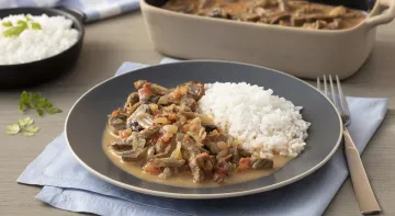 Fotografia em tons de cinza em uma bancada de madeira escura com um pano azul. Ao centro, um prato redondo preto com arroz e strogonoff de carne, ao lado um garfo e ao fundo uma tigela com strogonoff de carne e uma tigela com arroz.