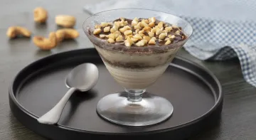 Fotografia em tons de marrom em uma bancada de madeira escura com um paninho azul xadrez. Ao centro, um prato de madeira redondo com uma taça de vidro e o pavê crocante com chocolate dentro dela. Ao fundo, castanhas-de-caju espalhadas.