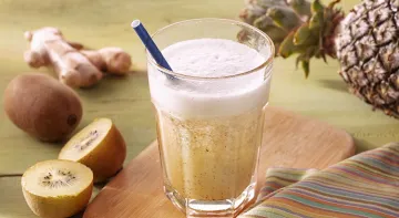 Fotografia em tons de amarelo em uma bancada de madeira de cor verde clara. Ao centro, uma tábua de madeira marrom com um copo de suco em cima. Ao lado, há 1 kiwi partido e ao fundo, outro kiwi, gengibre e um abacaxi. Fotografia em tons de amarelo em uma bancada de madeira de cor verde clara. Ao centro, uma tábua de madeira marrom com um copo de suco em cima. Ao lado, há 1 kiwi partido e ao fundo, outro kiwi, gengibre e um abacaxi.