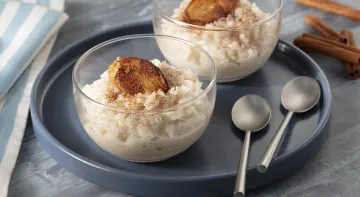 Fotografia em tons de cinza, em uma bancada de madeira escura com um pano azul listrado. Ao centro, um prato redondo preto, com duas taças de arroz doce com banana caramelizada e ao fundo, algumas canelas em pau espalhadas.