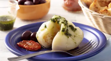 Fotografia em tons de branco em uma bancada de madeira de cor branca. Ao centro, um prato azul contendo a burrata com um garfo ao lado. Ao fundo, está uma cesta com pães e tomates secos.