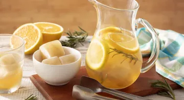 Fotografia em tons de laranja em uma bancada de madeira de cor marrom. Ao centro, um pano branco contendo uma tábua de madeira e em cima, há uma jarra contendo a água. Ao lado, cubos de gelo, um pegador e um pano quadriculado azul. Ao fundo, uma laranja.