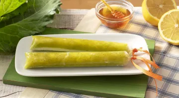 Fotografia em tons de verde em uma bancada de madeira de cor marrom. Ao centro, um pano branco quadriculado com um prato quadrado branco em cima contendo 2 geladinhos. Ao fundo, há uma couve, um pote de mel e dois limões.