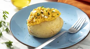 Fotografia em tons de amarelo em uma bancada de madeira de cor branca. Ao centro, um prato azul contendo a batata com um garfo ao lado. Ao fundo, há um pano laranja, um pote de azeite e algumas ervas espalhadas.