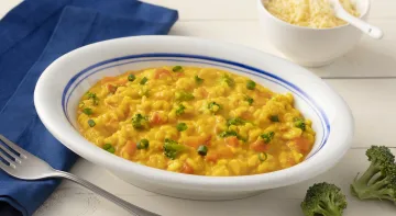 Fotografia em tons de azul em uma bancada de madeira clara com um pano azul escuro ao lado e um prato branco com detalhes em azul ao centro com o risoto de legumes com cúrcuma dentro. Ao lado, um potinho branco com queijo parmesão ralado e dois brócolis. Fotografia em tons de azul em uma bancada de madeira clara com um pano azul escuro ao lado e um prato branco com detalhes em azul ao centro com o risoto de legumes com cúrcuma dentro. Ao lado, um potinho branco com queijo parmesão ralado e dois brócolis.