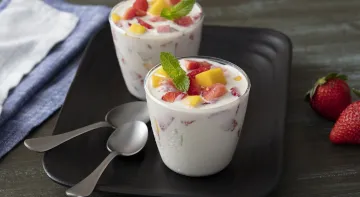 Fotografia em tons de azul, em uma bancada escura. Ao centro um prato quadrado preto com dois copos de vidro contendo a gelatina de frutas. Ao lado, dois morangos e um pano azul claro.