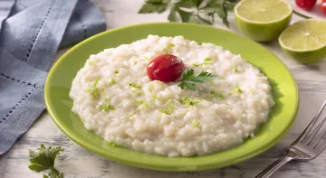 Fotografia em tons de verde em uma bancada de madeira de cor branca. Ao centro, um prato verde contendo o arroz. Ao lado, há um garfo e um pano azul. Ao fundo, há um limão partido ao meio, tomate cereja e salsinha espalhados. Fotografia em tons de verde em uma bancada de madeira de cor branca. Ao centro, um prato verde contendo o arroz. Ao lado, há um garfo e um pano azul. Ao fundo, há um limão partido ao meio, tomate cereja e salsinha espalhados.
