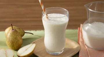 Fotografia em tons de branco em uma bancada de madeira de cor verde, ao centro, um pires de madeira contendo um copo com o smoothie. Ao lado, há algumas peras e do outro lado, um pano laranja quadriculado com uma jarra em cima. Fotografia em tons de branco em uma bancada de madeira de cor verde, ao centro, um pires de madeira contendo um copo com o smoothie. Ao lado, há algumas peras e do outro lado, um pano laranja quadriculado com uma jarra em cima.