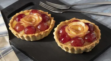 Fotografia em tons claros com uma toalha cinza listrada. Ao centro há um prato retangular preto com 2 tortas de frutas e ao fundo um par de garfos.