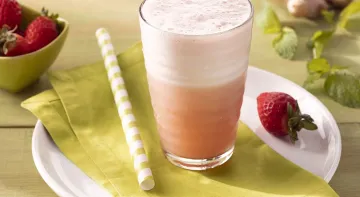 Fotografia em tons de laranja em uma bancada de madeira de cor verde clara. Ao centro, um prato branco com um pano verde em cima contendo um copo com a bebida. Ao lado, há um canudo e ao fundo, uma tigela com morangos, gengibre e folhas de hortelã.