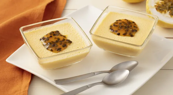 Fotografia em tons de laranja em uma bancada de madeira clara com um pano laranja, ao centro um prato branco quadrado e duas taças de vidro com o flan de maracujá dentro, enfeitados com sementes de maracujá. Ao fundo, um maracujá cortado ao meio. Fotografia em tons de laranja em uma bancada de madeira clara com um pano laranja, ao centro um prato branco quadrado e duas taças de vidro com o flan de maracujá dentro, enfeitados com sementes de maracujá. Ao fundo, um maracujá cortado ao meio.