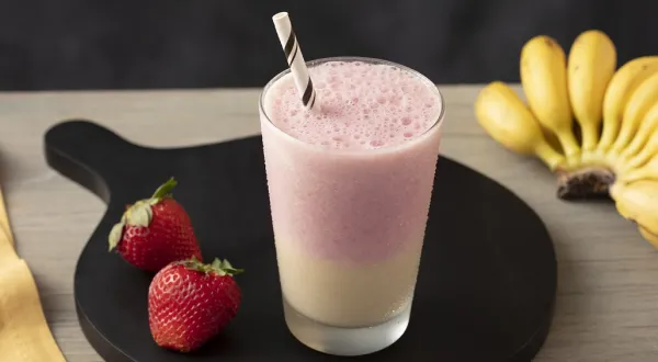 Fotografia em tons claros, em uma bancada de madeira bege escuro e ao lado, um pano amarelo. Ao centro, um prato redondo preto com um copo de smoothie morango e banana e um par de morangos ao lado. Ao fundo, um cacho de bananas. Fotografia em tons claros, em uma bancada de madeira bege escuro e ao lado, um pano amarelo. Ao centro, um prato redondo preto com um copo de smoothie morango e banana e um par de morangos ao lado. Ao fundo, um cacho de bananas.