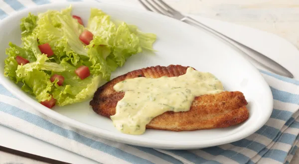Fotografia em tons de azul em uma bancada de madeira clara com um pano listrado azul claro e um prato oval branco com o filé de pescada com molho de limão e mostrada e salada de alface com tomates. Fotografia em tons de azul em uma bancada de madeira clara com um pano listrado azul claro e um prato oval branco com o filé de pescada com molho de limão e mostrada e salada de alface com tomates.