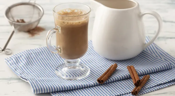 Fotografia em tons de azul em uma bancada de madeira clara com um pano azul com listras finas e ao centro uma taça com o cappuccino e ao lado uma jarrinha branca. Ao lado, três paus de canela e um difusor com o chocolate em pó. Fotografia em tons de azul em uma bancada de madeira clara com um pano azul com listras finas e ao centro uma taça com o cappuccino e ao lado uma jarrinha branca. Ao lado, três paus de canela e um difusor com o chocolate em pó.